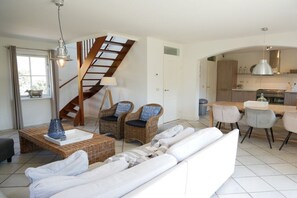 Ein modernes Wohnzimmer mit einer Couch, Korbstühlen, einem Esstisch und einer Küche im Hintergrund.
