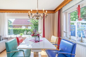 A dining area with a white table, blue and red chairs, a chandelier, and a window with curtains. - Holiday home with 3 bedrooms (Langenhorn)