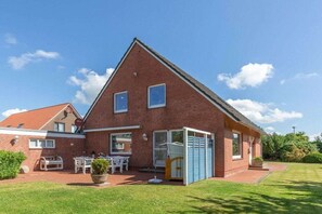 Ein rotes Backsteinhaus mit geneigtem Dach, einem umzäunten Garten und einer kleinen Terrasse mit Gartenmöbeln.