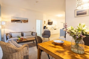 Ein modernes Wohnzimmer mit einer Couch, einem Couchtisch und einem Esstisch. Im Hintergrund ist eine Küche mit Schränken und einer Mikrowelle zu sehen.