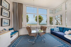 Ein Zimmer mit einem Glastisch, Korbstühlen, einem blauen Sofa und großen Fenstern mit Blick auf Bäume.