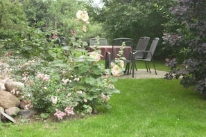 Ein Garten mit Tisch und Stühlen, umgeben von üppigem Grün und blühenden Pflanzen.
