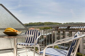 Ein Balkon mit zwei gestreiften Stühlen, einem kleinen Tisch mit Obstschale und Blick auf ein Gebäude sowie Bäume im Hintergrund.