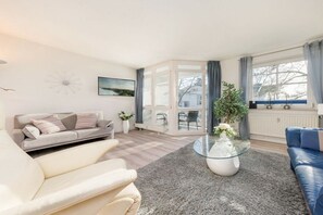 Uma sala de estar moderna com uma mesa de centro de vidro, um sofá, uma poltrona azul e vista para o exterior por uma porta de vidro.