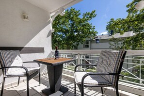 Ein Balkon mit zwei Korbstühlen, einem kleinen Tisch, einem weißen Geländer und Blick auf Bäume und Gebäude.
