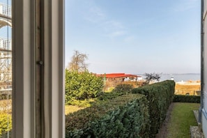 A view from a window overlooking a garden with a hedge, a building with a red roof, and a body of water in the distance. - Sea greetings at Villa Agnes (Binz)