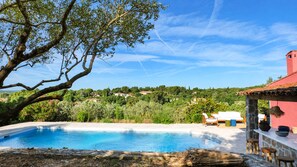 A swimming pool with a view of a hillside, a patio area with a table and chairs, and a red-roofed building. - Maison Olivette: Provençal villa with a private pool, 6 bedrooms, and olive trees (Flayosc)