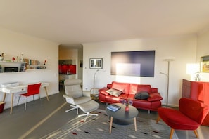 Ein modernes Wohnzimmer mit einem roten Sofa, einem weißen Sessel, einem kleinen Couchtisch und einem Schreibtisch mit Stuhl.
