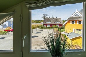 Blick durch ein Fenster auf ein Gebäude mit einem Reetdach und einer gelben Fassade, vor dem sich eine gepflasterte Fläche befindet.