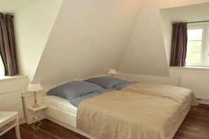 Un dormitorio con techo inclinado, una cama con cubrecama color beige, dos almohadas, una mesita de noche con lámpara y una ventana con cortinas.
