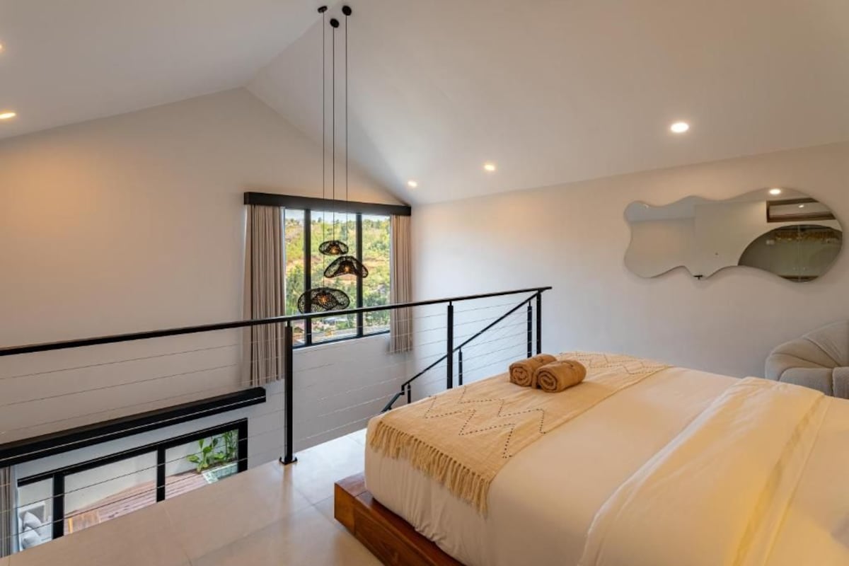 A modern bedroom with a large bed, a view of greenery through a window, a staircase, and a unique wall mirror.