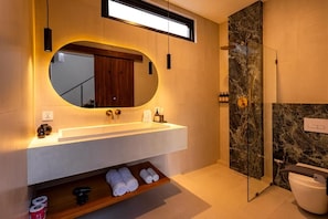 A modern bathroom with a large mirror, a built-in sink, a shower, and a shelf with towels. - Eala Estate (Pujut, Central Lombok Regency)