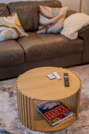 Ein runder Couchtisch aus Holz mit Fernbedienung und Buch, vor einer braunen Ledersofa mit gemusterten Kissen platziert.