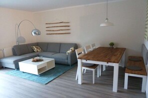 A modern living room with a grey sofa, a white coffee table, a wooden dining table with chairs, and a hanging lamp. - 14° EAST Holiday Apartment 1 (Koserow)