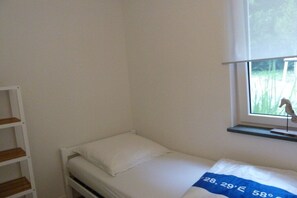 Une petite chambre avec un lit simple, une fenêtre, un lit blanc et une étagère en bois.