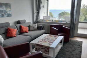 Een woonkamer met een grijze bank, een rode fauteuil en een witte salontafel met een tafelkleed met bloemenprint. Een raam biedt uitzicht op de zee.