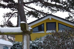 A yellow building with a triangular roof, surrounded by snow-covered trees and bushes. - Holiday home in Travemünde with Wi-Fi (Travemünde)