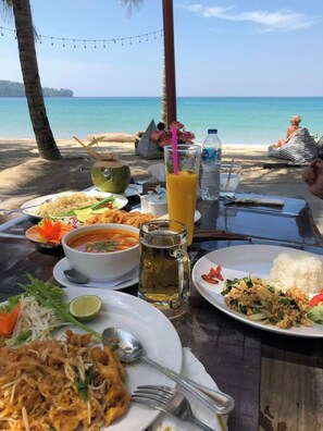 Een tafel gedekt met verschillende gerechten, een drankje en een kokosnoot op een strand, met uitzicht op de oceaan.