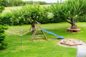 Ein Spielplatz mit Rutsche, Klettergerüst und Schaukeln in einer grünen Parkanlage mit Bäumen.