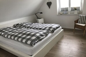 A bedroom with a bed, a nightstand, a chair, and a window with blinds. - House on the sea dike (Hedwigenkoog)