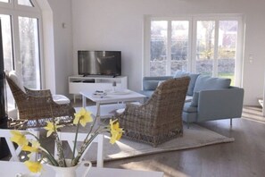 A modern living room with a television, wicker chairs, a sofa, and a vase of flowers. - House on the sea dike (Hedwigenkoog)