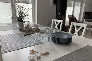 A dining table set with clear glassware, plates, and decorative items, with a potted plant and a fireplace in the background. - House on the sea dike (Hedwigenkoog)