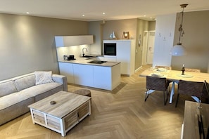 A modern kitchen with a built-in island, a dining area with a table and chairs, and a living space with a sofa and coffee table. - Holiday apartment with 1 bedroom (Lübeck-Travemünde)