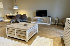 Een moderne woonkamer met een houten salontafel, een flatscreen televisie op een witte tv-meubel en een eethoek met stoelen en een tafel.