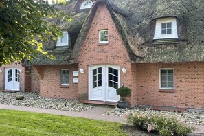 Ein traditionelles Backsteinhaus mit einem Reetdach, einem zentralen Eingang mit einer weißen Tür und einem kleinen Fenster darüber.