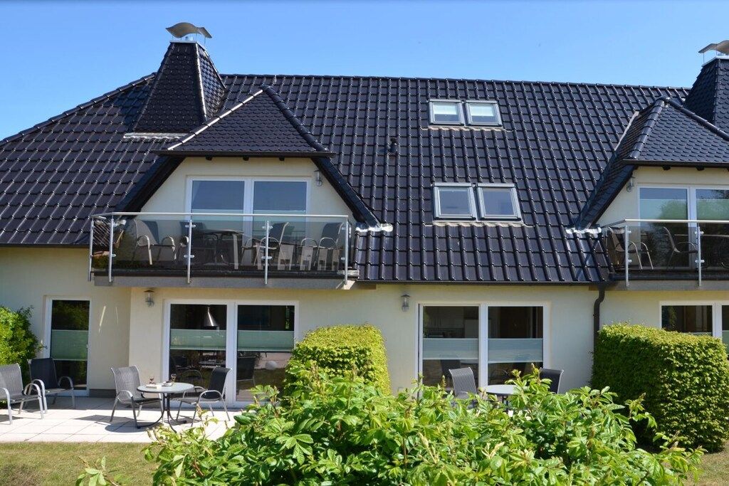 A two-story house with a dark tiled roof, large windows, and a balcony with outdoor furniture.