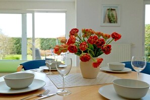 A dining table with white plates, bowls, and wine glasses, a vase of red flowers, and a view of a green landscape through large windows. - 3-room holiday apartment with terrace (Zinnowitz)