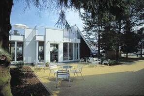 Un edificio moderno con tejado plano y ventanas amplias, rodeado de un patio empedrado con mesas al aire libre.