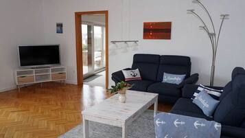 Ein modernes Wohnzimmer mit Fernseher, einer dunklen Sofagarnitur, einem weißen Couchtisch und einer Zimmerpflanze.