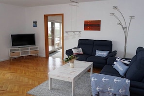 Ein modernes Wohnzimmer mit Fernseher, einer dunklen Sofagarnitur, einem weißen Couchtisch und einer Zimmerpflanze.