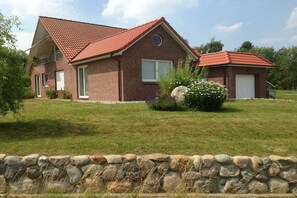 Ein zweistöckiges Backsteinhhaus mit rotem Ziegeldach, eine Steinmauer im Vordergrund und eine Rasenfläche.
