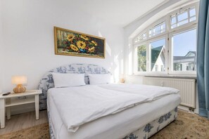 A bedroom with a bed, a nightstand, a lamp, a painting, and a window with a view. - “Meerweib” in Villa Sirene (Ostseebad Binz)