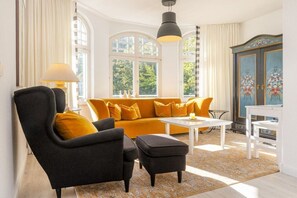 A room with a sofa, armchair, ottoman, and a table. There are windows with curtains, a lamp, and a cabinet with decorative elements. - “Meerweib” in Villa Sirene (Ostseebad Binz)