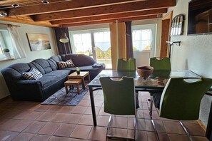Un salon chaleureux avec un canapé, une table en verre et des chaises. Il y a une fenêtre avec des rideaux, un tableau au mur et un plafond en bois.