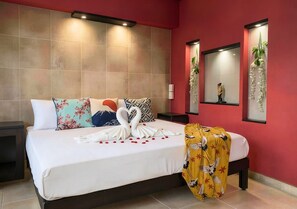 Habitación de hotel con una cama grande, almohadones decorativos, un espejo montado en la pared y una planta en la mesita de noche.
