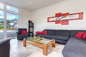 Un salon moderne avec une table basse en verre, des canapés gris et des coussins rouges. On aperçoit une cheminée dans un coin.