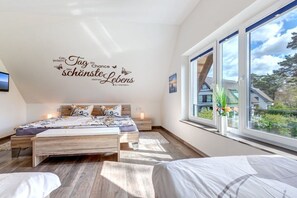 Ein Schlafzimmer mit großem Fenster, einem Bett mit gemustertem Überwurf, einem Nachttisch mit Lampe und einer Wanddekoration mit Text.