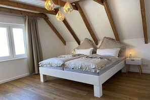 Ein Schlafzimmer mit schrägen Wänden, einem Bett mit weißem Gestell, einem Nachttisch mit Lampe und einem Fenster mit Vorhängen.