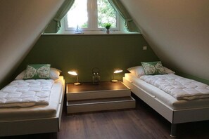 Ein Zimmer mit zwei Betten, jeweils mit Kissen und einer Nachttischlampe. Ein Fenster mit Vorhängen und einem Blumentopf auf der Fensterbank.