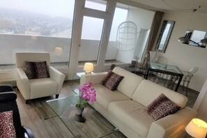 Ein modernes Wohnzimmer mit Sofa, Sessel, Glastisch und Blick auf die Stadt durch große Fenster.