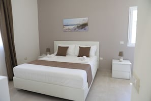 Ein ordentlich bezogenes Bett mit weißen Textilien und braunen Akzenten, ein Nachttisch mit Lampe und ein Bild an der Wand.