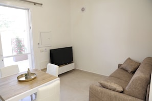 Ein Wohnzimmer mit Flachbildfernseher auf einem Schrank, einer Couch, einem Esstisch mit Stühlen und einem Fenster mit Blick auf Grünflächen.
