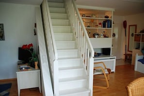 Escales que condueixen a un segon pis, una taula consola blanca amb un televisor i objectes decoratius, i una prestatgeria plena de llibres i objectes decoratius.