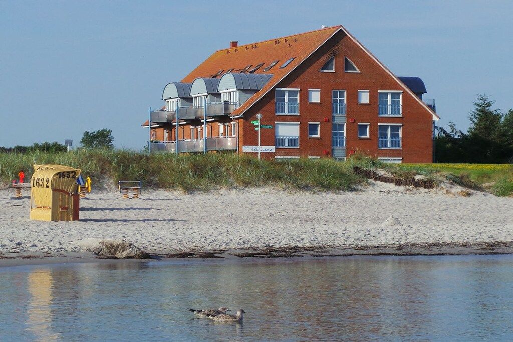A red brick building with a sloped roof, a water body in the foreground, and a sandy beach.