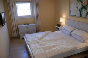 Un dormitori amb un llit, una tauleta de nit, una làmpada, una finestra amb cortines i un quadre a la paret.