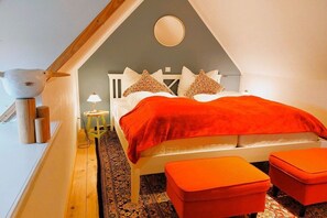 A bedroom with a sloped ceiling, a bed with a red blanket, two matching ottomans, a small stool, and a round mirror on the wall. - Holiday home with 2 bedrooms (Steinbergkirche)